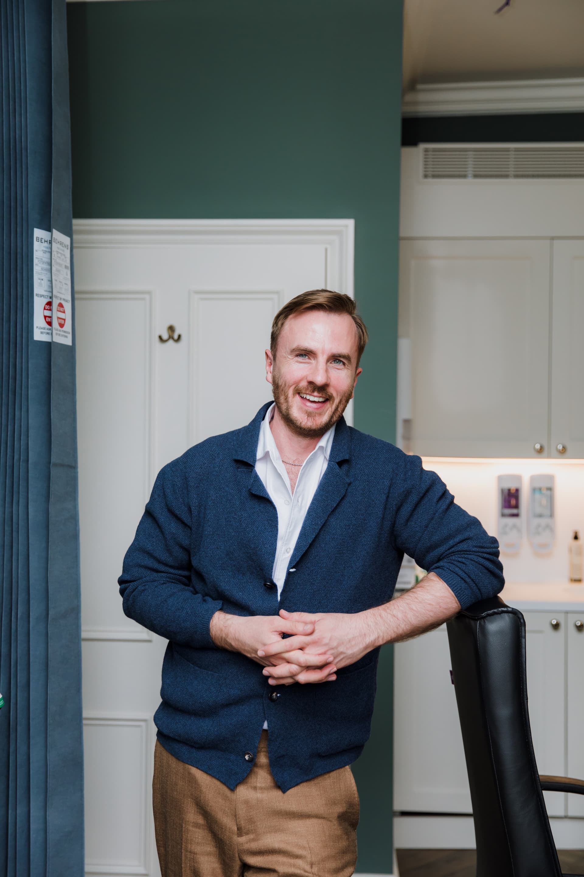 Dr Ben Davis standing beside a black leather chair with his hands loosely clasped, in a navy cardigan over an open shirt, smiling, in a teal-green room at the practice